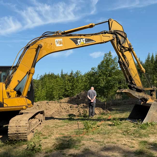 Excavator Building a home is a creative act. A large yellow CAT excavator frames a man standing with his hands behind his back as he looks over a freshly excavated home site marked with orange survey flags beneath a blue summer sky.